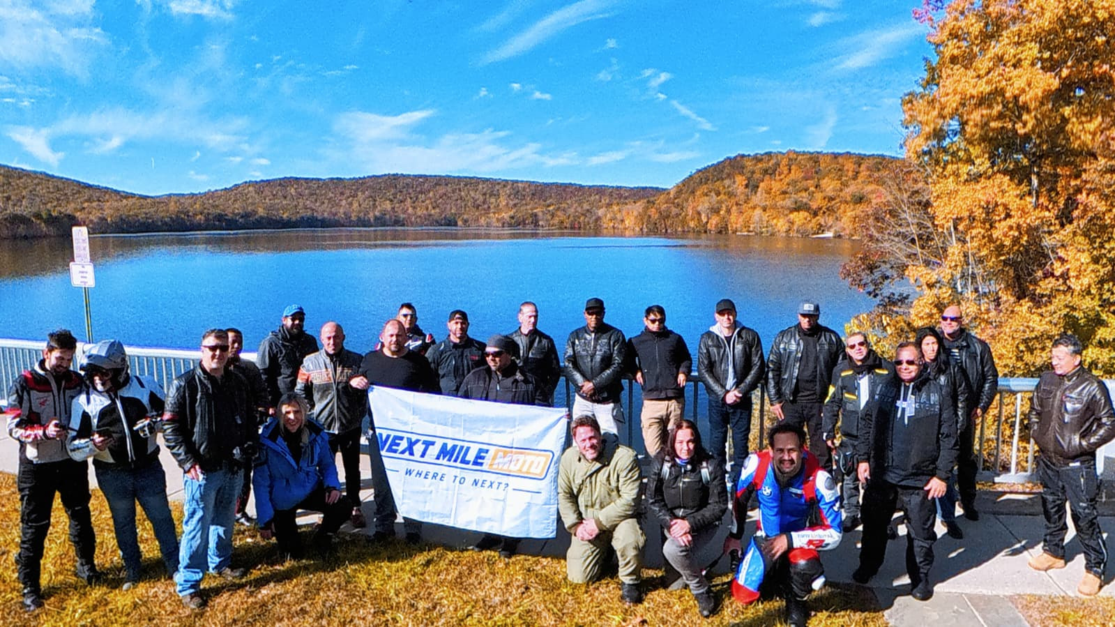 Next Mile Moto riders gathered with the group banner beside a fall reservoir during The Autumn Mile, October 24, 2026